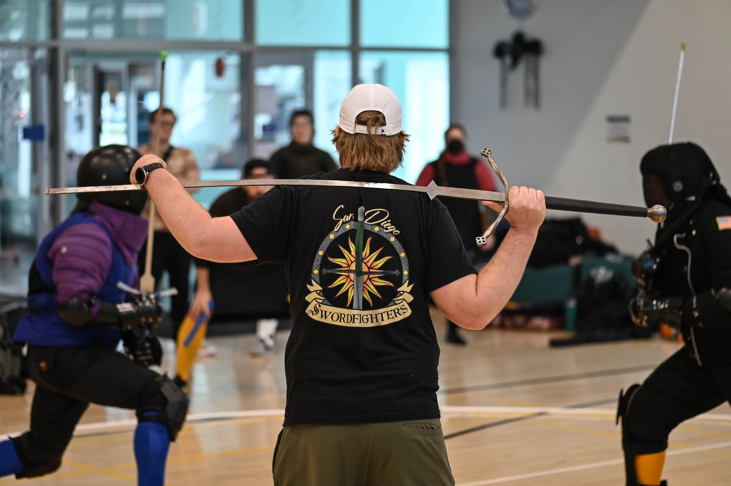 People practicing wooden sword combat in an indoor gym, with spectators in the background.
